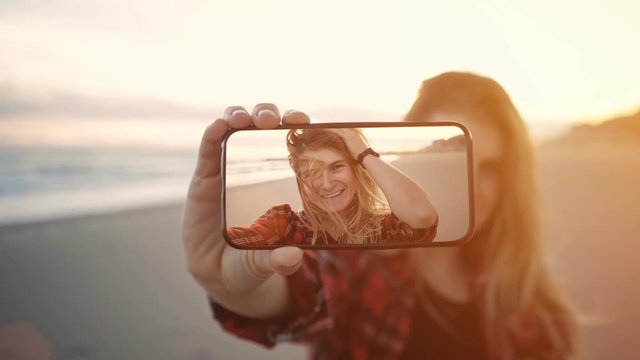 Cinemagraph of young teenage girl or student woman with blond hair and in red pleaded shirt smile and laugh, makes selfies during sunset at beach on summer vacation