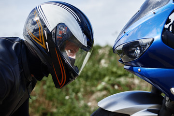 Close up sideways view of blue motorcycle and self determined young male rider wearing black leather jacket and stylish protective helmet getting ready for race. Hobby, and active sports concept