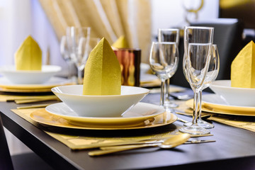 Set table with golden cutlery and napkins, white and golden dishes, different glasses, golden tablecloths and black table. The napkins are on the upper plates as decoration. Golden wall