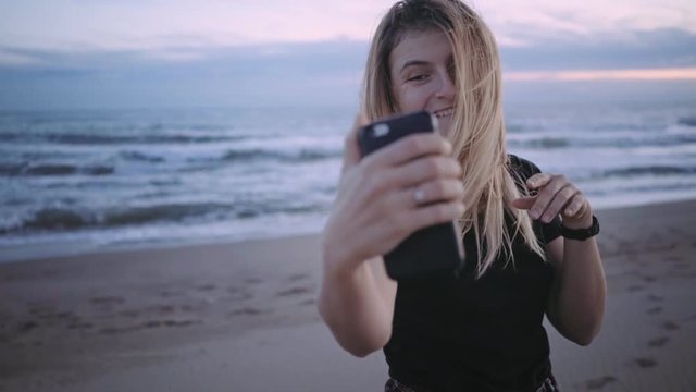 Slow motion video of attractive young millennial hipster blonde girl or woman at beach,excited and happy,laughs and smiles while makes funny faces to selfie on smartphone camera,poses for social media