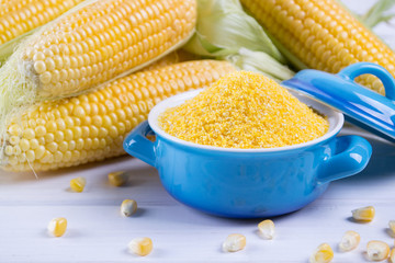 Corn grits polenta in a blue ceramic bowl on white table with ripe raw corn cob and green leaves