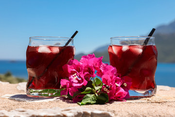 Two glasses of summer red cocktail with ice and a sprig of Bougainvillea flowers between on the blue sky background, summer sea vacation concept.