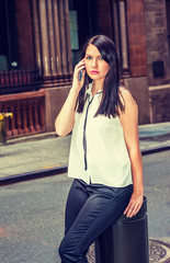 Fototapeta premium Young American Woman with long black hair, wearing white collarless sleeveless shirt, black pants, sitting on vintage narrow street in New York, listening, talking on cell phone. Filtered effect.