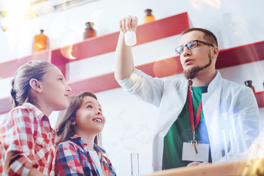 Please Turn Your Attention To The Jar. Male Up And Coming Scientist Wearing A Laboratory Coat Showing A Glass Jar With A Chemical Liquid His Amazed Little Pupils Standing Next To Him In The Lab.