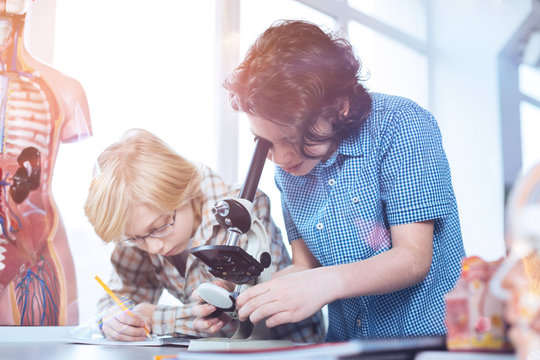 Closer Look. Young Persistent Active Boys Working On Their Experimental Assignment While One Of Them Using A Microscope And The Other Writing Down Data