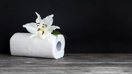 Paper towel roll - paper tissue and lily flower on table against black background. Body care and hygienic disposables concept. Copy space.