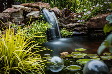 A lush koi pond surrounded by stones and plants with a misty waterfall