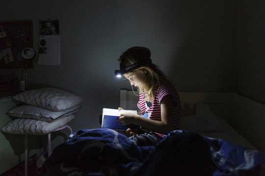 Young girl reading book with head torch while sitting in bed at night