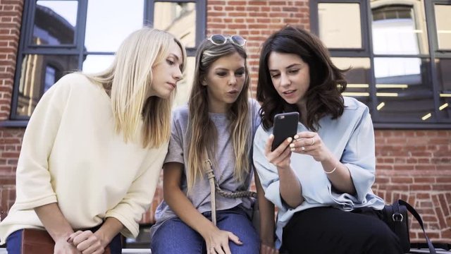 Three young women looking at a smartphone screen while sitting in the summer street. Zoom out real time medium shot