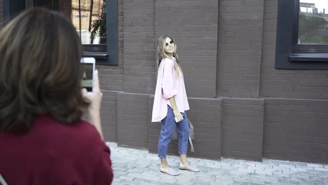 Unrecognizable dark haired woman making shots of a blonde model wearing a pink shirt and glasses in a city street. Handheld real time medium shot