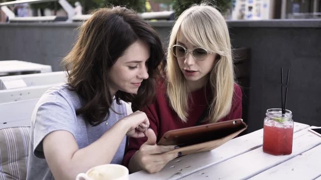 Two girlfriends are sitting in an outdoors cafe on a summer day and web surfing from a digital tablet. Zoom out real time medium shot