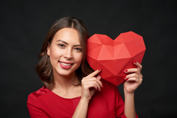 Closeup of smiling woman in red holding red polygonal paper heart shape over dark background