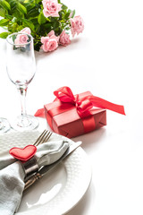 Valentines day dinner with table place setting with red gift, glass for champagne, heart ornaments with silverware on white. Close up. Valentine's card.