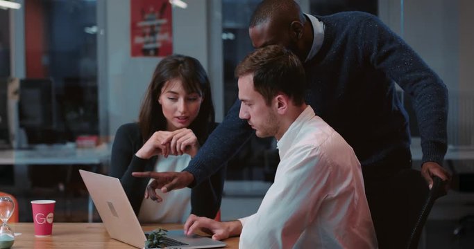Employees of prestigious law firm preparing important project on Friday evening. Employees attentively looking at laptop screen, discussing project, making suggestions. Indoors.