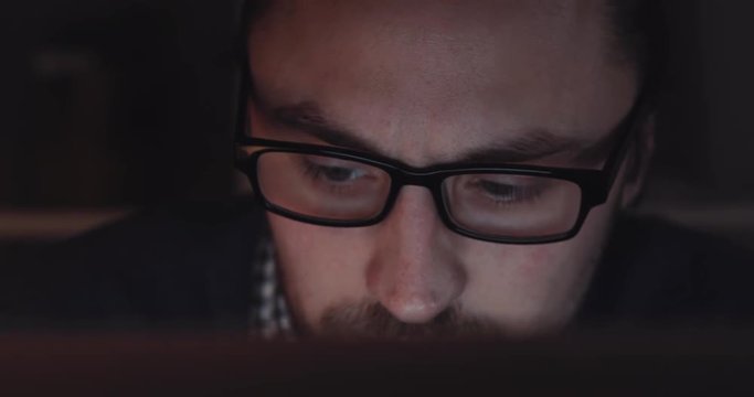 Handsome young man in spectacles working hard at some important project on the laptop late at night. Workaholic, being successful. Close up view