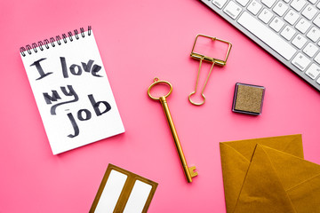 Happy at work. Handwritten motto I love my job in notebook near stationery on pink background top view