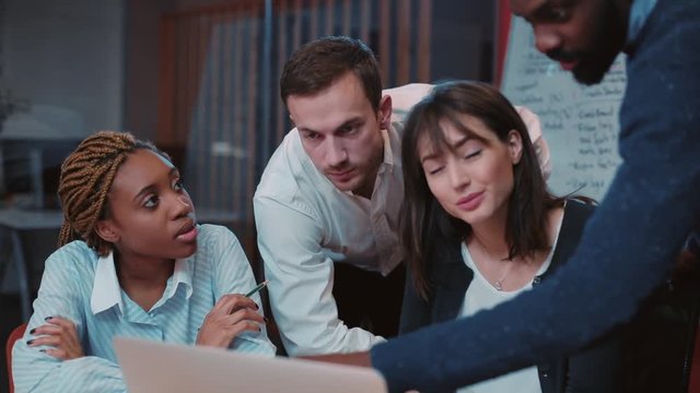 Young African man demonstrating prepared presentation in beautiful office. Serious young people listening attentively to new employee. Guy pointing at board.