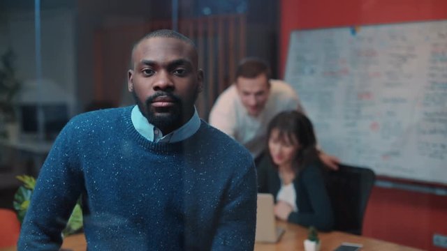 Young serious African man with beard in blue sweater looking directly at camera on background of modern office. Young people working, looking at monitor.