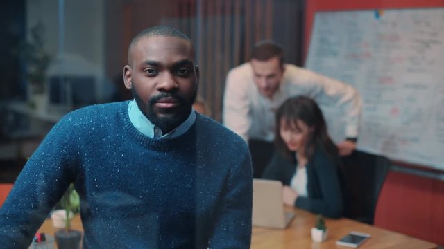 Likable young African man in blue sweater with beard looking at camera and smiling. Employees of office working on background. Evening. Indoors.