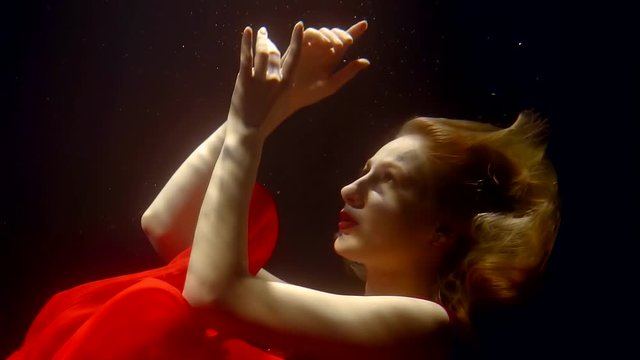 extravagant art dancer woman is moving under water in darkness, she is waving hands, red dress