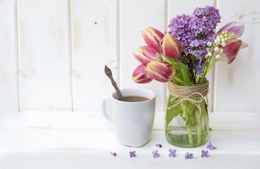 cup of coffee with milk and bouquet of tulips and lilac on white wooden background