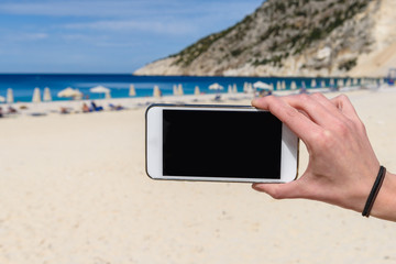 Women's hand holding blank screen smartphone or mobile on a beach. Vacation, travel concept.