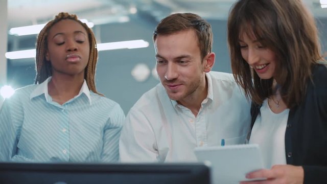 Young people of different nationalities standing near computer and looking at monitor, discussing something in beautiful modern office. Smiling. Indoors.