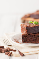 Chocolate brownie square pieces in stack on white plate decorated with mint leaves and cocoa powder on white vintage wooden background. American traditional delicious dessert. Close up photography