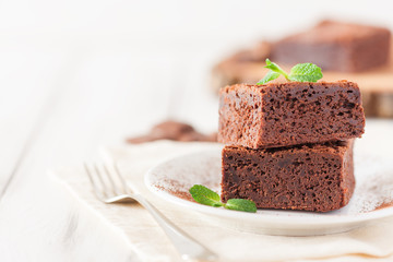 Chocolate brownie square pieces in stack on white plate decorated with mint leaves and cocoa powder on white vintage wooden background. American traditional delicious dessert. Close up photography