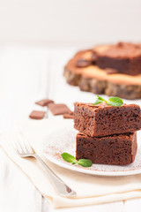 Chocolate brownie square pieces in stack on white plate decorated with mint leaves and cocoa powder on white vintage wooden background. American traditional delicious dessert. Close up photography
