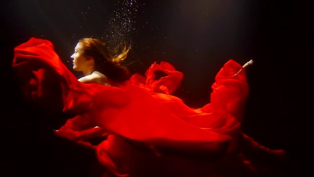 mystical redhead undine is floating underwater with red fabric around in dark deepness of river