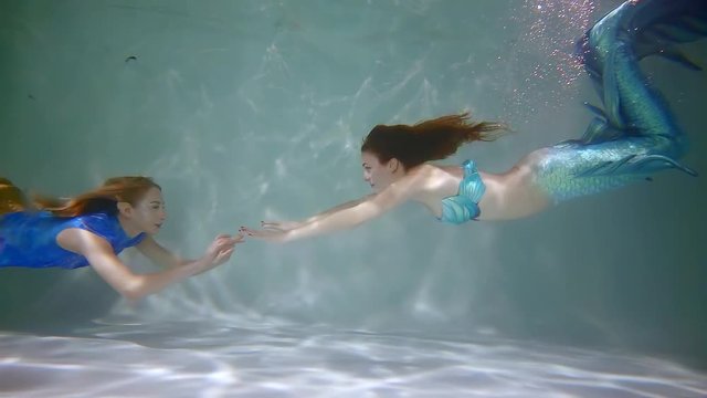 two fairy mermaid are floating underwater in light clear pond, sunshine is brighting through water