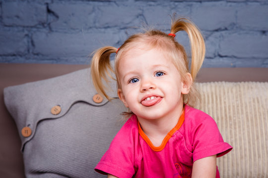 Portrait Of A Little Funny Blonde Girl With Blue Eyes And A Funny Hairdo Dabbles And Plays On The Sofa At Home. He Laughs Showing His Tongue And Teeth. She Is Dressed In Bright Stylish Clothes