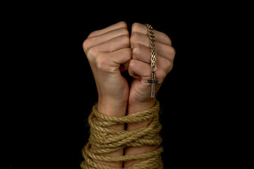 hands tied with a rope holds a religious symbol a silver cross on a chain  close-up black background