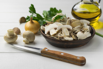 Fresh sliced mushrooms in a clay bowl and onion and oil on a white wooden table.