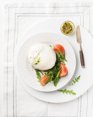 Italian cheese Burrata (mozzarella) with fresh arugula, tomatoes, olive oil, sauce pesto on a white plate and a wooden light background. Flat lay, top view