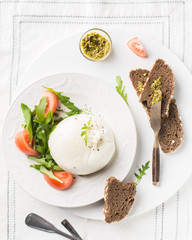 Italian cheese Burrata (mozzarella) with fresh arugula, tomatoes, olive oil, sauce pesto and whole grain bread toasts on a white plate and a wooden light background. Flat lay, top view