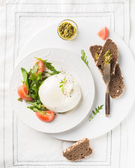 Italian cheese Burrata (mozzarella) with fresh arugula, tomatoes, olive oil, sauce pesto and whole grain bread toasts on a white plate and a wooden light background. Flat lay, top view