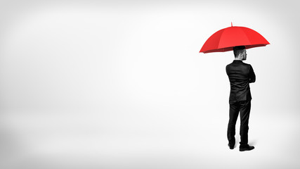 A high contrast view on a businessman standing in half-turn under an open red umbrella.