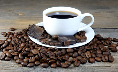 White cup of coffee with a bunch of coffee beans and pieces of chocolate on a wooden table