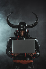 armored samurai warrior using laptop on dark background with smoke