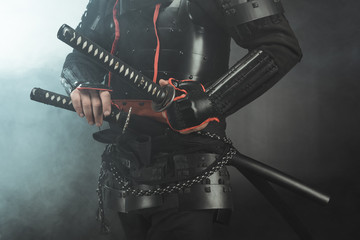 cropped shot of samurai in armor with swords on dark background with smoke