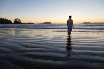 Fototapeta premium Woman Walking on a Sandy Beach during a vibrant and colorful summer sunset. Taken in Tofino, Vancouver Island, BC, Canada.