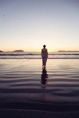 Fototapeta premium Woman Walking on a Sandy Beach during a vibrant and colorful summer sunset. Taken in Tofino, Vancouver Island, BC, Canada.