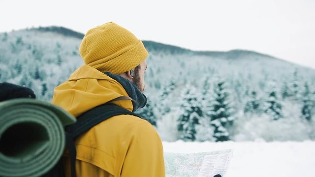 Beautiful mountains in winter time. A guy with beard, wearing yellow winter clothes is wandering and holding a map in his hands, against the background of the winter landscape