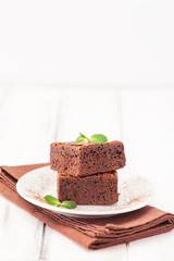 Chocolate brownie square pieces in stack on white plate decorated with mint leaves and cocoa powder on white vintage wooden background. American traditional delicious dessert. Close up photography