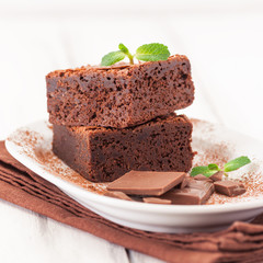 Chocolate brownie square pieces in stack on white plate decorated with mint leaves and cocoa powder on white vintage wooden background. American traditional delicious dessert. Close up photography