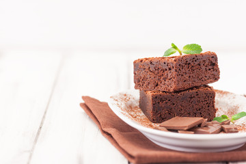 Chocolate brownie square pieces in stack on white plate decorated with mint leaves and cocoa powder on white vintage wooden background. American traditional delicious dessert. Close up photography