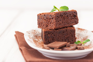 Chocolate brownie square pieces in stack on white plate decorated with mint leaves and cocoa powder on white vintage wooden background. American traditional delicious dessert. Close up photography