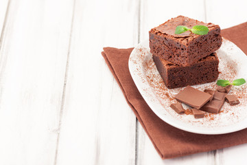 Chocolate brownie square pieces in stack on white plate decorated with mint leaves and cocoa powder on white vintage wooden background. American traditional delicious dessert. Close up photography
